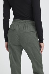 Šedé joggingové kalhoty s elastickým pasem a dvěma zadními kapsami. Tkanina má texturovaný, měkký vzhled. Viditelná dlouhá černá košile.