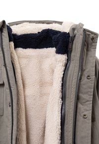 Chaqueta aislante en verde oliva, con forro de borrego crema y cuello azul marino. Cuenta con cierre frontal de cremallera y botones a presión.