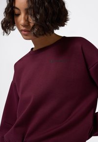 Mujer con cabello corto y rizado, vistiendo una sudadera burdeos de Champion, mirando hacia abajo contra un fondo blanco liso.
