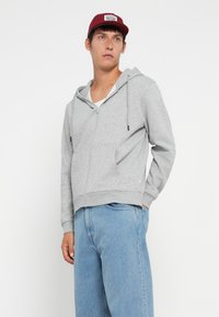 Grauer Reißverschluss-Hoodie mit einer Fronttasche, verstellbarem Kordelzug an der Kapuze und gerippten Bündchen, kombiniert mit hellblauen Jeans und einer weinroten Mütze.
