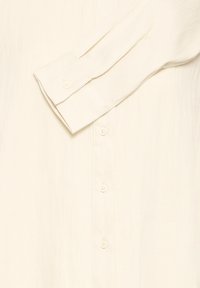 Long-sleeve beige button-up cotton shirt with cuffs and white buttons shown flat on a light background.