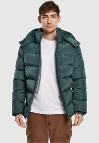Tyrkysová péřová bunda se zapínáním na zip a kapucí. Má prošívaný design a elastické manžety; nošena přes bílý tričko a hnědé kalhoty.