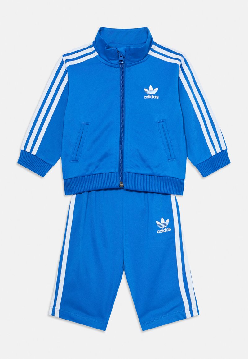 Blå idrottsdräkt i mjukt tyg, med en zip-up jacka med vita ränder och logotyp, matchande shorts med elastisk midja.