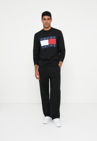 Sudadera negra con un logo frontal en rojo, blanco y azul. Combinada con pantalones negros de corte relajado y zapatillas blancas.