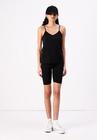 Top senza maniche nero con spalline sottili e pantaloni shorts in bici neri abbinati, indossati con sneaker alte bianche e un cappellino nero.