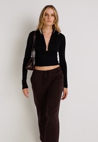Melna garām piedurknēm iesaiņota blūze, saskaņota ar brūnām augstā vidukļa sporta biksēm, kas izceļas ar teksturētu audumu un minimālistisku dizainu.