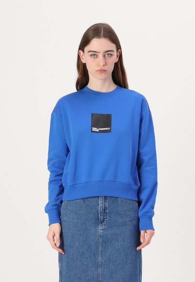 Sweatshirt bleu royal avec un col rond et des poignets côtelés. Présente un logo noir en forme de carré sur la poitrine. Associé à une jupe en denim.