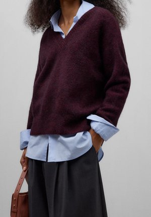 Personne portant un pull col V bordeaux superposé à une chemise bleu clair avec les manches retroussées, jupe plissée foncée, tenant un sac à main marron.