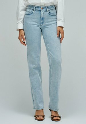 Femme portant un jean bleu clair à jambes droites, taille haute avec fermeture à boutons, assorti à des sandales marron à lanières et un chemisier blanc à manches longues.