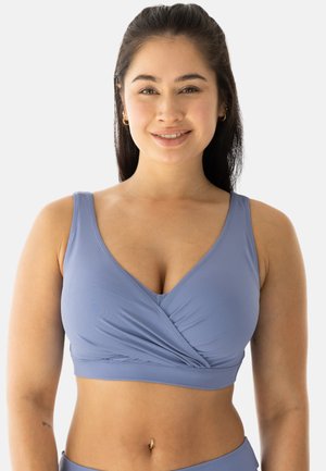 Young woman with long dark hair wears light blue sports bra and gold hoop earrings, smiling against a plain white background.