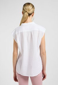 Blusa blanca de manga corta en tejido texturizado, con un cuello clásico, pliegue en la parte posterior y dobladillo curvado. Combinada con pantalones rosas claros.