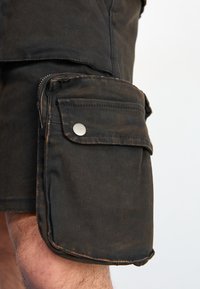 Calções de carga pretos feitos de algodão resistente, com um bolso lateral de remendo, botão de pressão prateado e uma textura gasta e desbotada.
