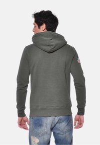 Haut à zip couleur vert foncé avec une grande capuche, des poignets côtelés et une taille côtelée. Présente un petit écusson du drapeau américain sur le bras supérieur gauche.