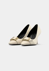 Cream high-heeled shoes with a pointed toe, featuring a gold buckle accent and black interior. Smooth texture with a structured design.