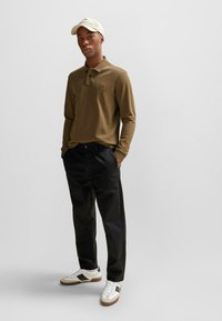 Bruine lange mouwen polo met kraag, zwarte broek en witte sneakers met donkere accenten. Een lichtgekleurde pet maakt de outfit compleet.