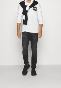 Langärmliges weißes T-Shirt mit einem schwarzen Logo, kombiniert mit dunklen Jeans und schwarzen Sneakers. Ein schwarz-weißes Sweatshirt liegt über den Schultern.