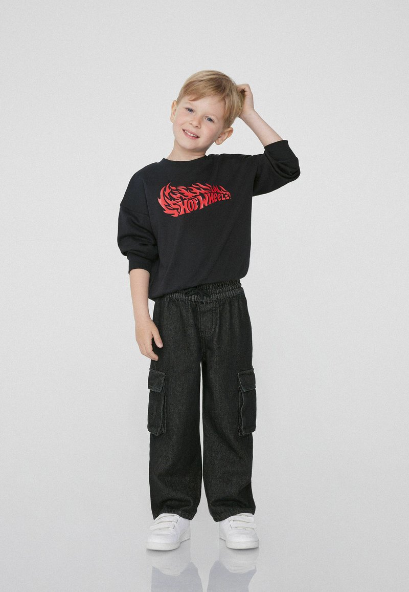 Czarna bluza z kapturem z czerwonym napisem "Hot Wheels", zestawiona z luźnymi, ciemnoniebieskimi spodniami cargo i białymi trampkami.