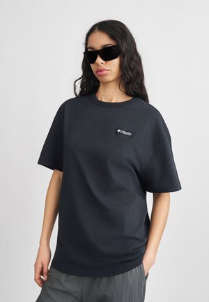 Woman wearing black oversized Columbia t-shirt and dark sunglasses, standing with one hand in pocket against plain background.