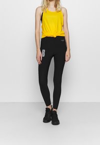 Kobieta w żółtej bluzce bez rękawów zawiązanej w talii, czarnych legginsach z nadrukowanym literą "B" i czarnych sportowych butach stoi na tle białej ściany.