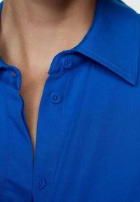 Camicia button-up di un blu brillante, con colletto strutturato, due bottoni blu e una texture del tessuto liscia. Vista ravvicinata del colletto.