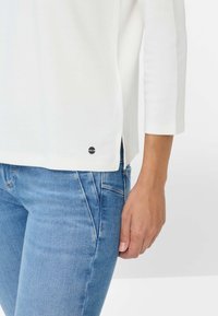 Weißes Langarmoberteil mit seitlichen Schlitzen und kleinem schwarzem Logo am Saum, kombiniert mit hellblauen, eng anliegenden Jeans. Glatte Textur, lässiges Design.