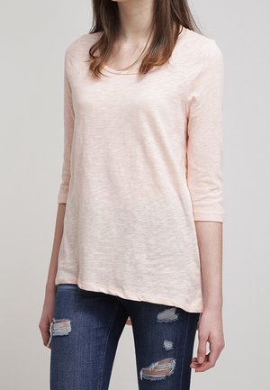 Kobieta ubrana w jasnoróżowy top z rękawem do łokcia i podarte ciemnoniebieskie dżinsy, stojąca na jednolitym tle.