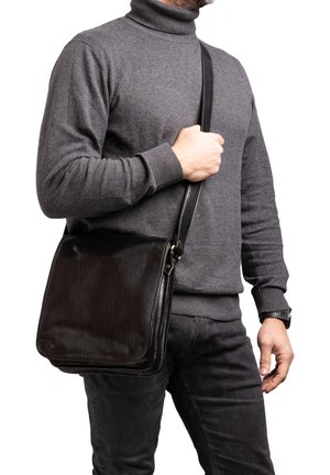 Man in een grijze coltrui en zwarte spijkerbroek houdt een zwarte leren schoudertas vast aan het hengsel, over de borst en taille.