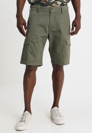 Homme portant un short cargo vert olive avec des poches latérales, associé à des baskets blanches, debout sur un fond blanc.