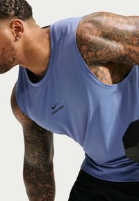 Μυώδης άνδρας με μπλε αμάνικο μπλουζάκι Nike Pro σηκώνει μια μαύρη αλτήρα, δείχνοντας λεπτομερή τατουάζ στα χέρια και στο λαιμό σε απλό φόντο.