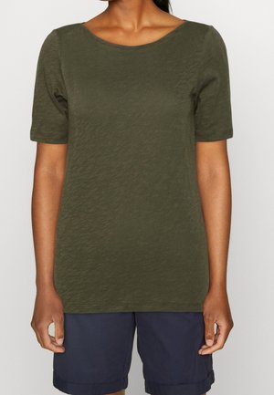 Personne portant une chemise à manches courtes vert olive et un short bleu marine, debout devant un fond clair uni.