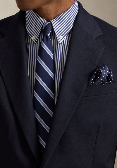 Blazer azul-marinho sobre uma camisa de fato riscada azul e branca, com uma gravata às riscas azul e branca e um lenço de bolso azul-marinho com bolinhas brancas.