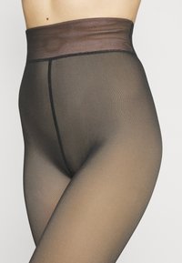 Genomskinliga svarta strumpbyxor med en hög, slät midja. Materialet har en fin, texturerad yta och sömmarna är snyggt sydda med svarta trådar.
