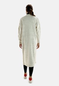 Krämfärgad lång poncho i ribbad textur med flätstickade detaljer längs sidorna, bärs över svarta leggings och vita sneakers med röda accenter.