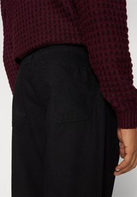 Burgundy neulepaita, jossa on ruutukuvio, yhdistetty mustiin housuihin, joissa on taskuyksityiskohtia ja sileä kangas.