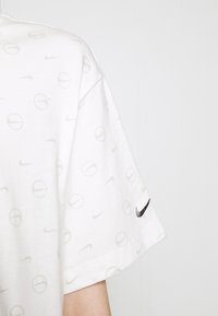 Bílé bavlněné tričko s jemnými šedými logy Nike a kruhovými vzory, s tmavým logem Nike na levém rukávu.