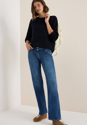 Maglione blu navy a trama con maniche lunghe abbinato a jeans dritti azzurri e sneakers marroni. Lo sfondo è di tono neutro.