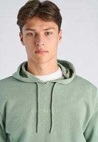Jonge man met lichtbruin haar, gekleed in een saliegroene hoodie met de tekst "LINDBERGH", kijkt direct in de camera tegen een effen achtergrond.