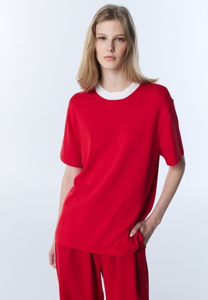 Giovane donna con capelli biondi lisci che indossa una maglia rossa a maniche corte con colletto bianco e pantaloni rossi abbinati, con la mano in tasca.