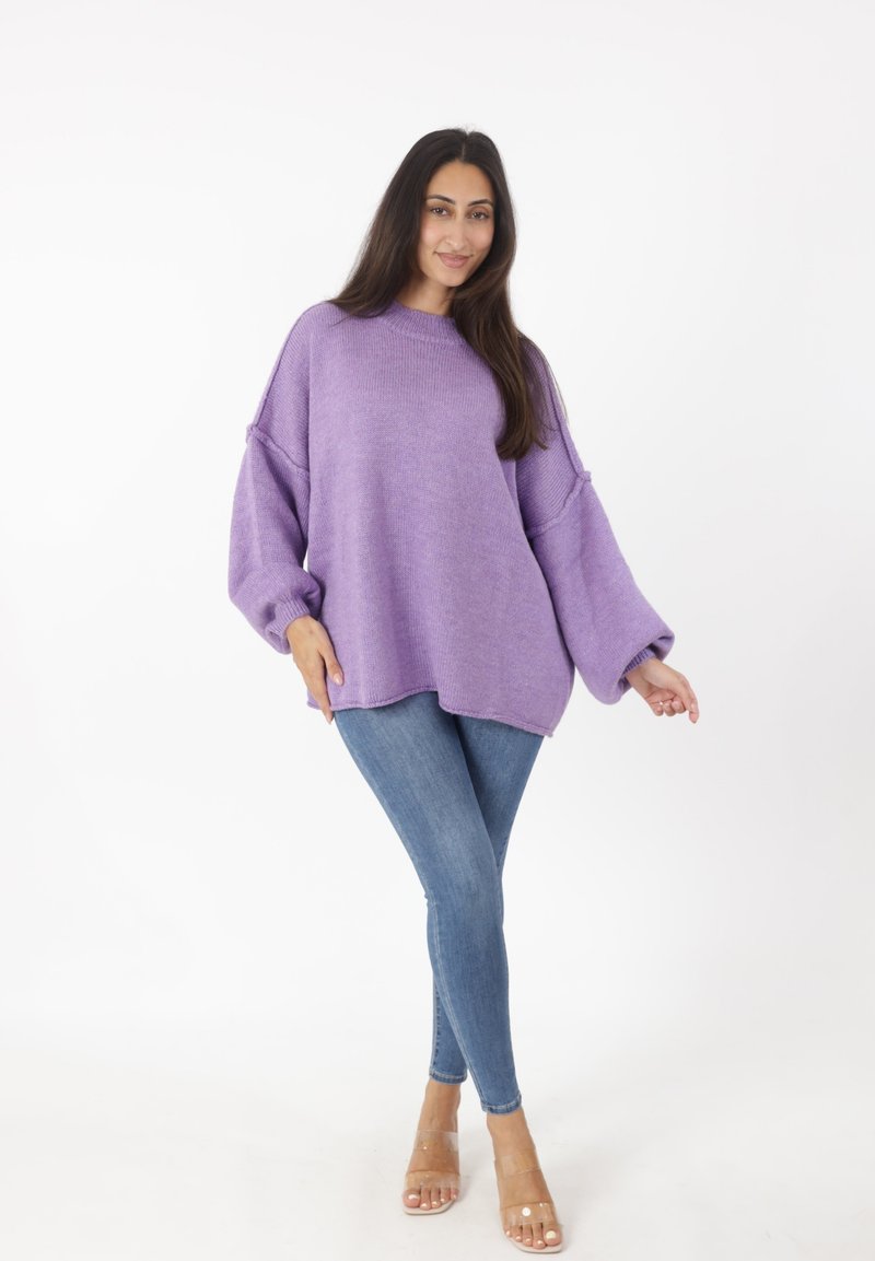 Lila übergroßer Strickpullover mit fallen Schultern, kombiniert mit engen blauen Jeans und klaren Absatzsandalen auf einem weißen Hintergrund.