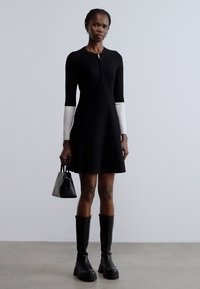 Čierne priliehavé šaty s dlhými bielymi rukávmi, textúrovaná látka, zapínanie na zips, doplnené malou čiernou kabelkou a čiernymi vysokými čižmami po kolená.