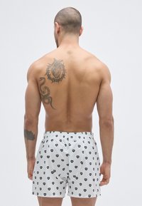 Boxer bianchi con motivo a cuori neri, vita elastica e tasche laterali. Indossati da un modello maschile senza maglietta e con un tatuaggio sulla schiena.
