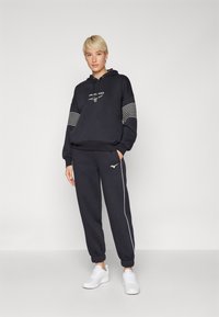 Navy hettegenser med grafisk logo, stripete detaljer på ermene, sammen med matchende joggebukser med hvit side stripe og logo. Fottøyet er hvite sneakers.