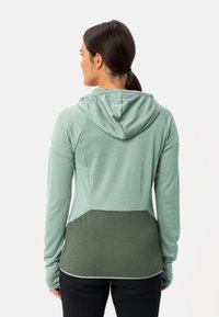 Giacca verde con cappuccio, maniche lunghe, tessuto texturizzato e orlo curvo. Pannello di un verde più scuro in contrasto nella parte inferiore.