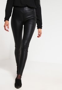 Zwarte faux leren leggings met een hoge taille, slim-fit ontwerp, gladde textuur en minimale stikseldetails, gecombineerd met zwarte schoenen.