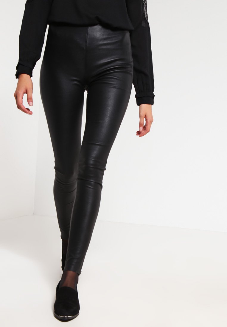 Zwarte faux leren leggings met een hoge taille, slim-fit ontwerp, gladde textuur en minimale stikseldetails, gecombineerd met zwarte schoenen.