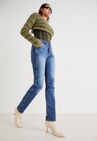 Suéter corto de punto en verde oliva, top oscuro, jeans azules de cintura alta y botines de tacón beige. La modelo posa con las manos en las caderas.