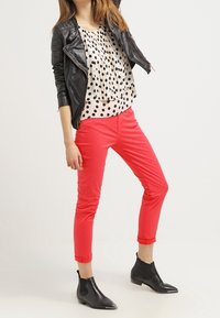 Giacca da biker in finta pelle nera, blusa bianca con pois e pantaloni aderenti corallo. Stivaletti neri alla caviglia completano il look.