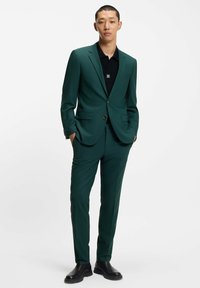 Completo verde a taglio slim con rever a punte, bottone singolo e tasche laterali. Indossato sopra una camicia nera, abbinato a stivali neri.