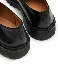 Scarpe di pelle nera con suole in gomma spessa e testurizzata. Forma arrotondata, finitura liscia e cucitura sul retro. L'interno è di colore beige.
