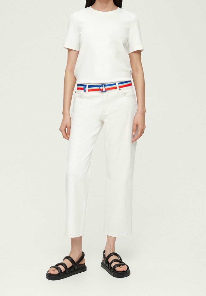 Femme portant un haut blanc à manches courtes, un pantalon blanc avec une ceinture rayée rouge, blanche et bleue, et des sandales noires à bouts ouverts.